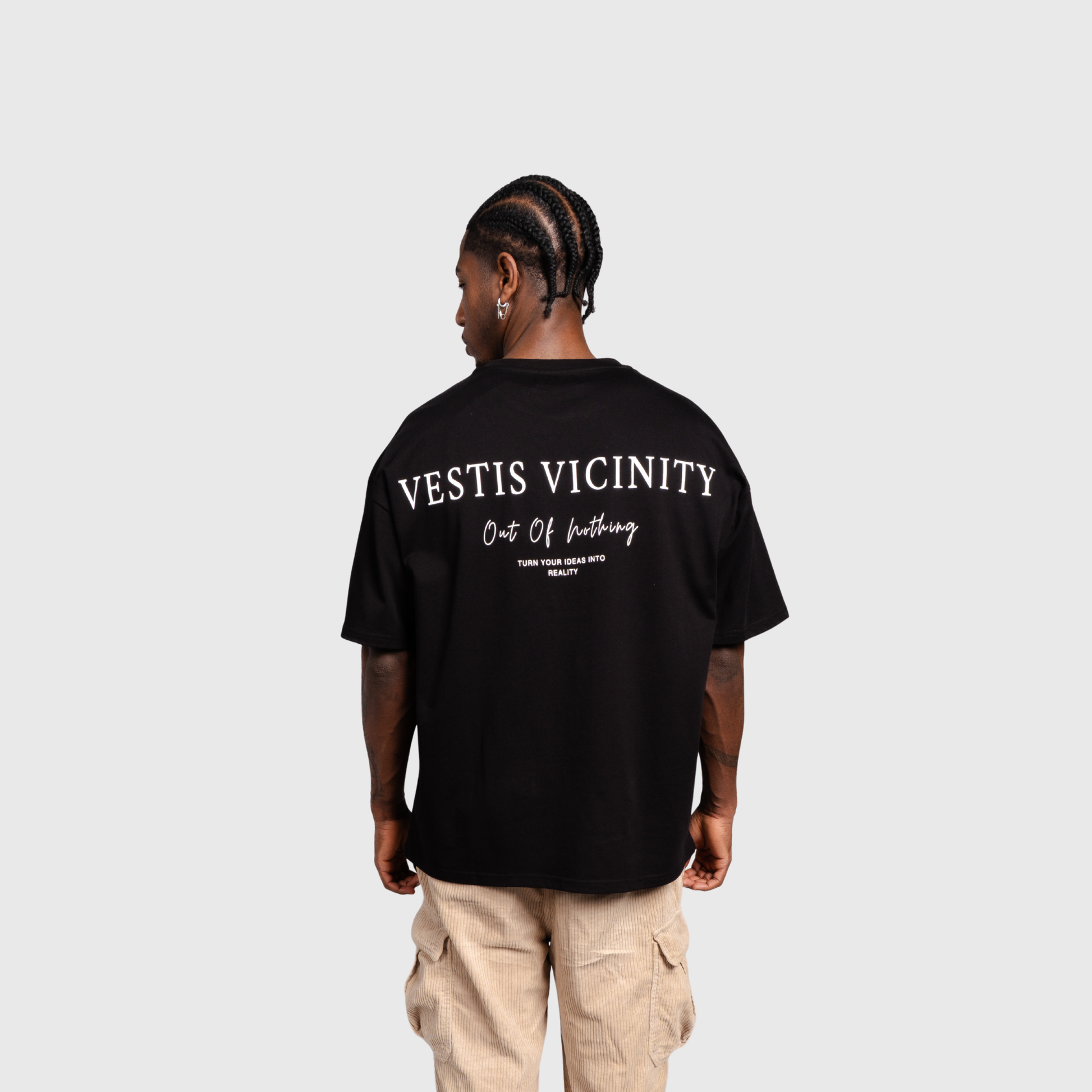 Model standing with their back to the camera wearing a black oversized T-shirt and beige cargo pants. Large white text graphic is printed across the upper back
