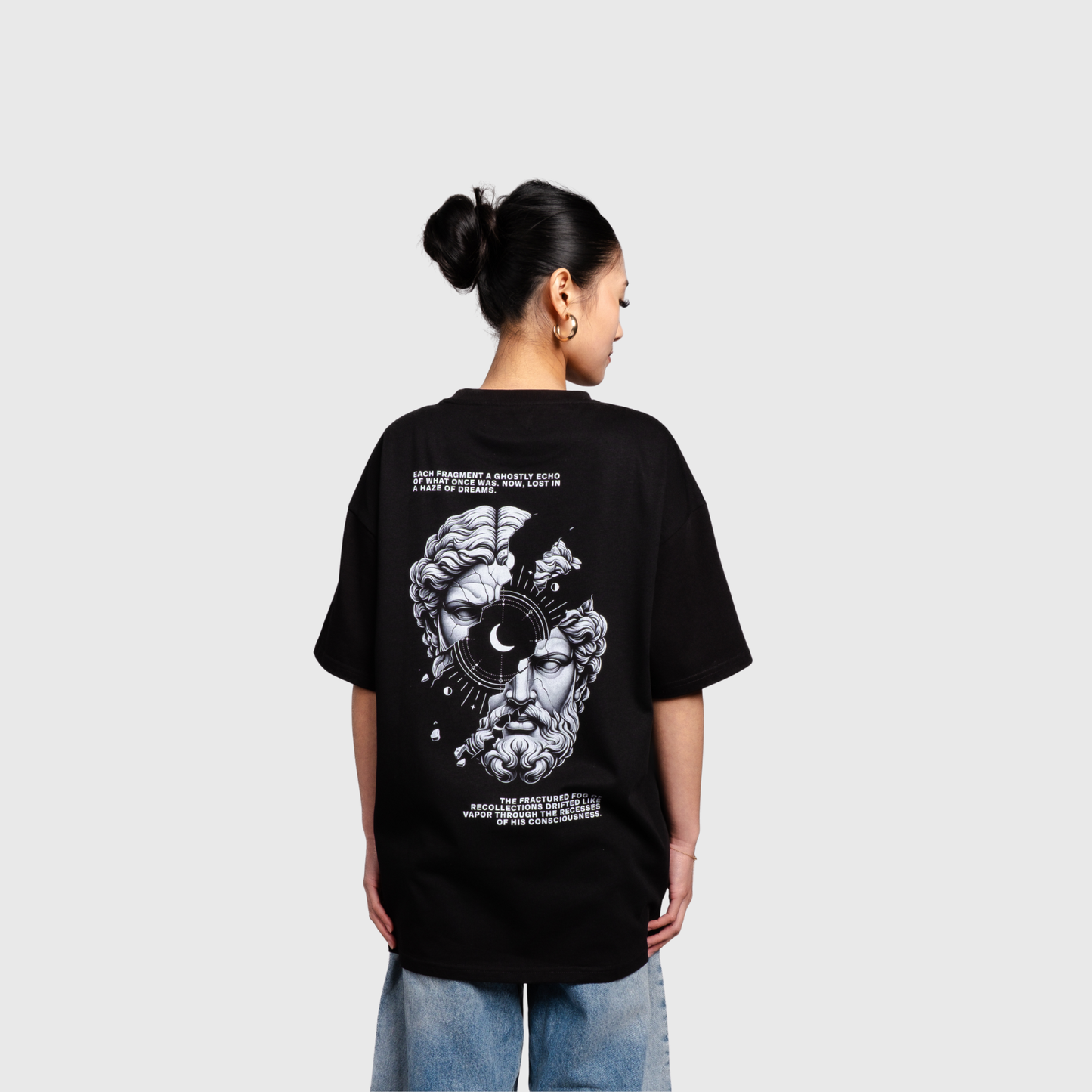 Model standing with their back to the camera wearing a black oversized T-shirt. The back features a large monochrome illustration of a classical statue face combined with abstract and celestial design elements