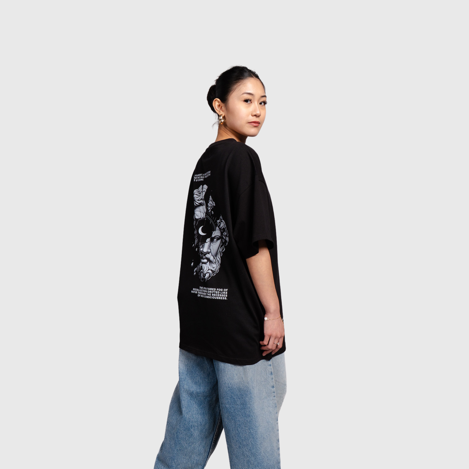 Side view of a model wearing a black oversized graphic T-shirt and light blue jeans. Part of the large back artwork is visible along the side of the shirt