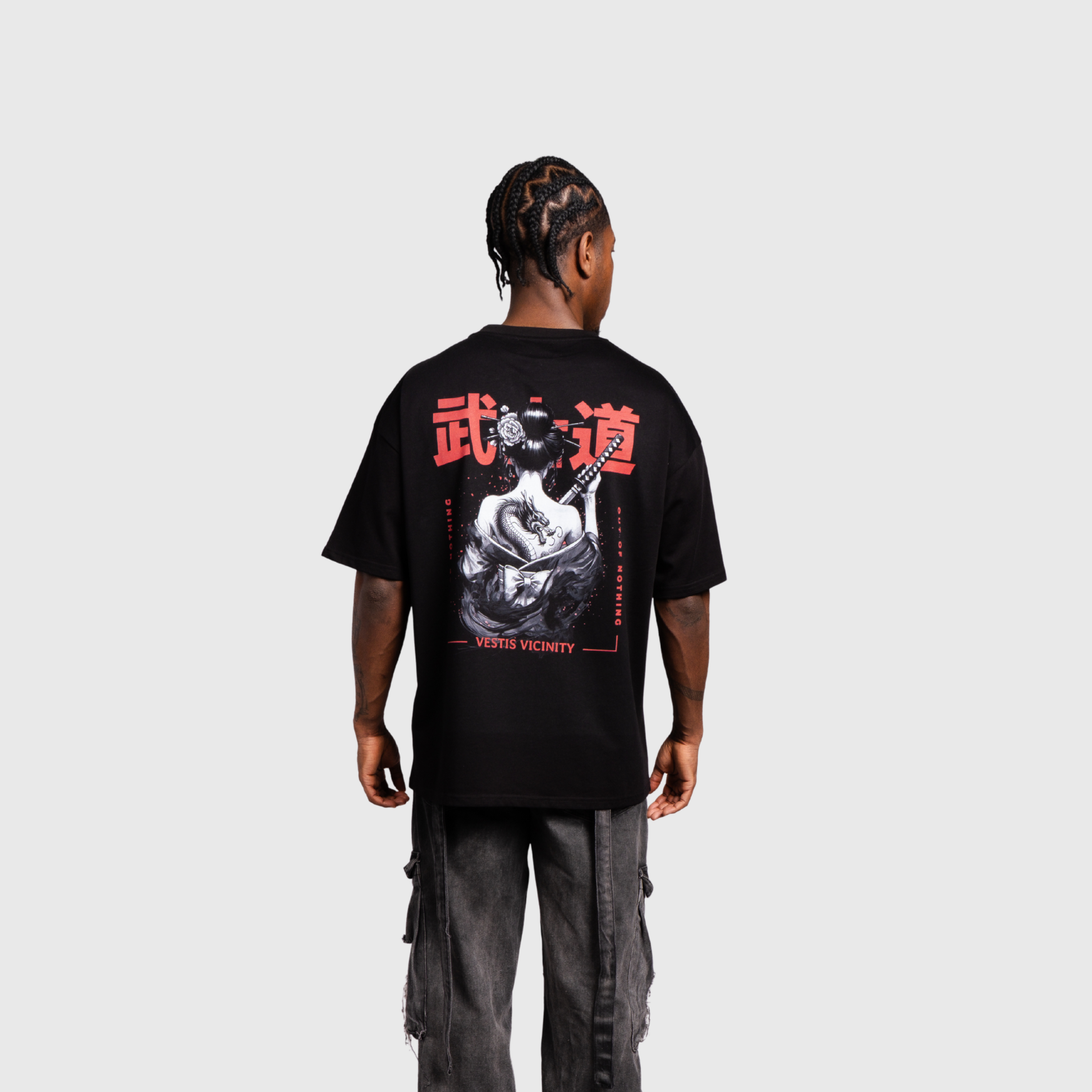 Model standing with their back to the camera wearing a black oversized graphic T-shirt. The large back print features a monochrome warrior artwork framed by bold red Japanese-style characters and graphic details.