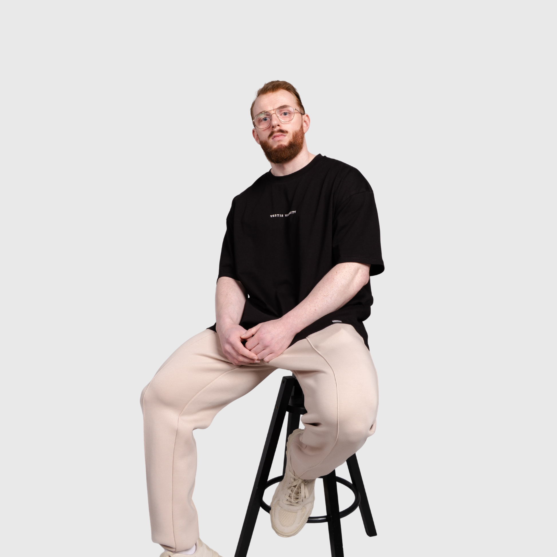 Model seated on a stool wearing a black oversized T-shirt, beige pants, and light-colored sneakers. The shirt features a small text graphic on the chest