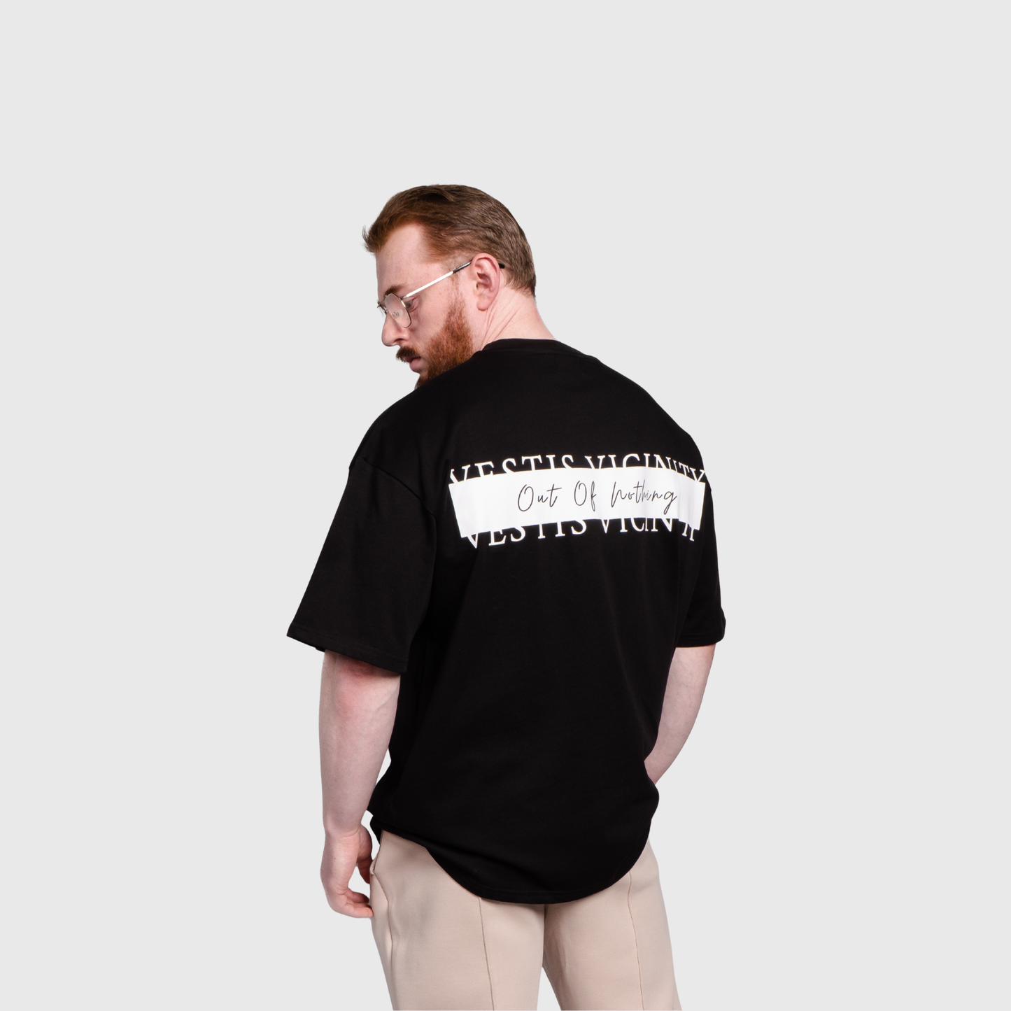 Model standing and looking downward while showing the back of a black oversized T-shirt. A large text graphic with a horizontal gray stripe spans across the upper back