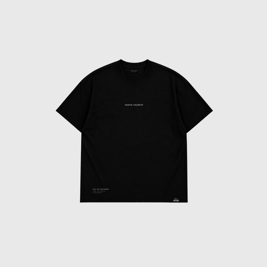 Front view of a black oversized T-shirt on a light gray background. Small minimalist text detail appears on the chest, with a subtle graphic near the lower hem